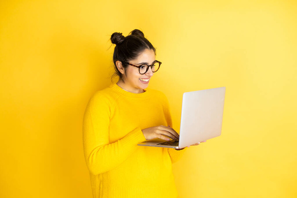 Młoda brunetka pracuje przy użyciu laptopa na żółtym tle ze szczęśliwą twarzą stojącą i uśmiechniętą z pewnym siebie uśmiechem pokazującym zęby - Zdjęcie, obraz