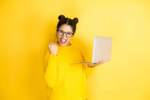 Fiatal barna nő dolgozik számítógép laptop sárga háttér nagyon boldog és izgatott, hogy a győztes gesztus emelt karokkal, mosolygós és sikoltozó siker. - Fotó, kép