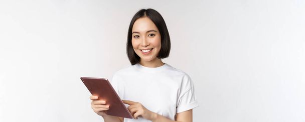 Asyalı kız dijital tablet kullanıyor, aletlerle çalışıyor, internetten alışveriş yapıyor, beyaz arka planda duruyor. - Fotoğraf, Görsel