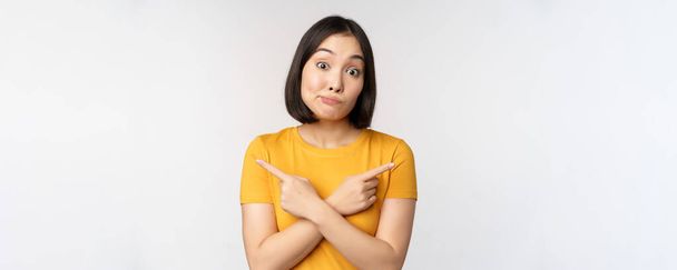Сложная азиатская девушка, указывающая пальцами в сторону, показывающая левый и правый выбор, глядя нерешительно невежественный, стоя на белом фоне - Фото, изображение