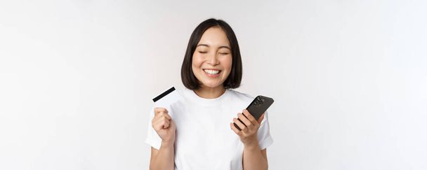 Интернет-магазины. Счастливая азиатка с помощью кредитной карты и смартфона приложение, оплата на сайте с помощью мобильного телефона, белый фон - Фото, изображение