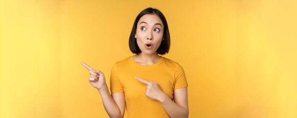 Portret zaintrygowanej Azjatki, patrzącej i wytykającej palcami w lewo na reklamę, pokazującej smth ciekawy, stojący nad żółtym tłem - Zdjęcie, obraz