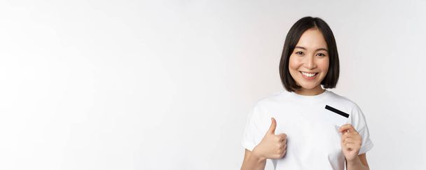 Retrato de hermosa joven mujer asiática moderna, mostrando tarjeta de crédito y pulgares hacia arriba, recomendando el pago sin contacto, de pie sobre fondo blanco - Foto, imagen
