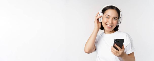 Beautiful modern asian girl, listening music in headphones, holding mobile phone, using smartphone app, standing over white background - Фото, зображення
