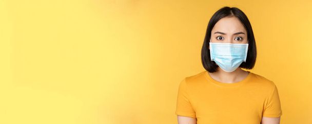 Muotokuva järkyttynyt aasialainen nainen näyttää huolissaan ja säikähti kameraa, yllään covid-19 lääketieteellinen kasvonaamio, seisoo keltaista taustaa vasten - Valokuva, kuva
