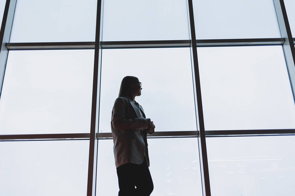 Een succesvolle zakenvrouw in broek en een jas staat en kijkt uit op de enorme panoramische raam van het business center wolkenkrabber. - Foto, afbeelding