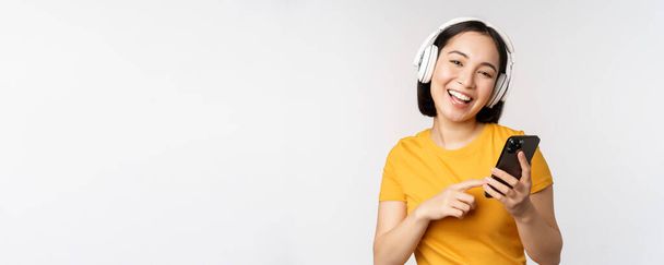 Cute japanese girl in headphones, looking at mobile phone and smiling, using music app on smartphone, standing against white background - Фото, изображение
