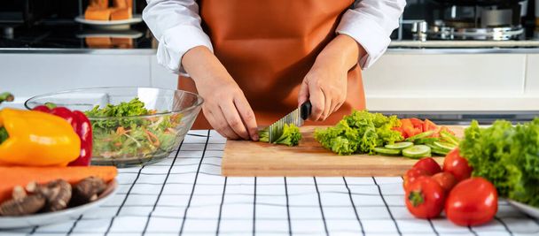 Azji gospodyni domowa za pomocą noża do krojenia sałaty na desce do krojenia drewna do przygotowania składników do sałatki warzywnej podczas noszenia fartuch i stojąc do gotowania zdrowego posiłku w kuchni w domu - Zdjęcie, obraz