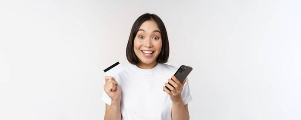 Compras en línea. Mujer asiática feliz usando la tarjeta de crédito y la aplicación de teléfono inteligente, pagando en el sitio web a través del teléfono móvil, fondo blanco - Foto, imagen