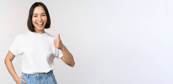Happy beautiful korean woman, smiling pleased, showing thumbs up in approval, recommending brand or company, standing over white background - Foto, Imagen