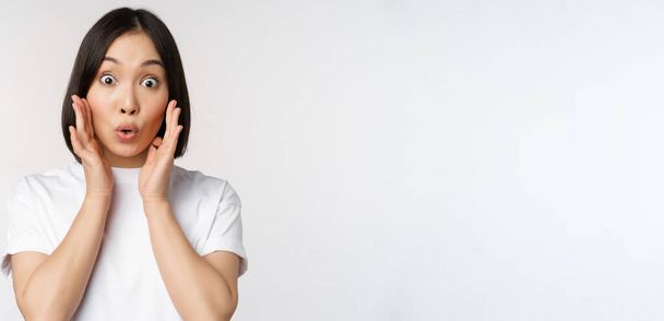 Close up portrait of asian girl looking surprised, wow face, reacting amazed at smth, standing in white tshirt over studio background, isolated - 写真・画像