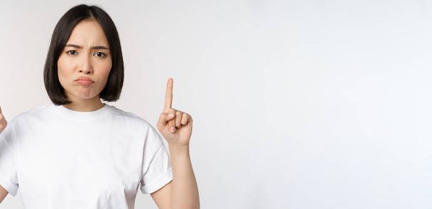 Sad and disappointed young asian woman pointing fingers up, showing advertisement with upset face expression, white studio background - Фото, зображення