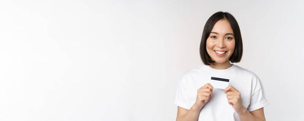 Улыбающаяся корейская женщина показывает кредитку со счастливым лицом, стоя в футболке на белом фоне - Фото, изображение