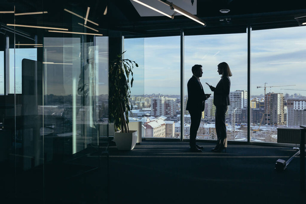 Két üzletember, ázsiai férfi és nő sziluettjei tanácskoznak és kommunikálnak, egy modern irodában dolgoznak az ablak mellett. - Fotó, kép