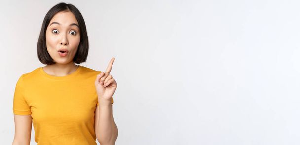 Beautiful young asian woman pointing finger up, smiling and looking amused at camera, showing advertisement, announcement on top, white background - Zdjęcie, obraz