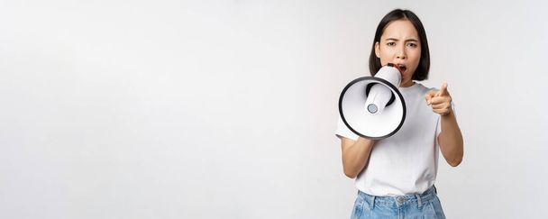 Angry asian woman with megaphone, scolding, accusing someone, protesting with speakerphone on protest, standing over white background - Фото, изображение