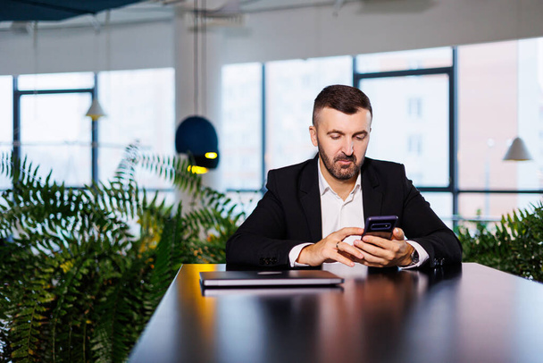 Hombre de negocios confiado en un traje de negocios se sienta en una mesa con un teléfono y trabaja remotamente - Foto, Imagen