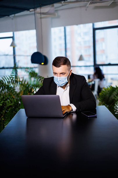 Adulto exitoso hombre de negocios guapo en una máscara con un ordenador portátil mientras trabaja en una oficina remota en un café - Foto, imagen