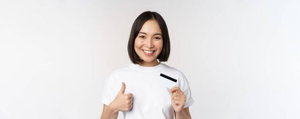 Güzel, genç, modern Asyalı bir kadının portresi, kredi kartı ve onaylar, temassız ödeme önerir, beyaz arka planda durur. - Fotoğraf, Görsel