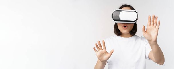 Amused asian girl using VR glasses, virtual reality headset and reaching hands into empty space, touching smth augmented, standing over white background - Photo, Image