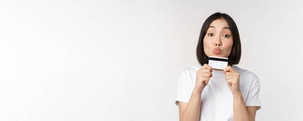 Money and finance concept. Cute japanese girl kissing her credit card, standing in tshirt over white background - Photo, Image