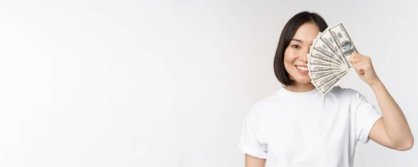 Портрет усміхненої азіатки, що тримає долари гроші, концепція мікрокредитування, фінанси та готівки, стоїть на білому тлі
 - Фото, зображення