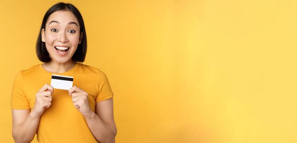 Beautiful asian woman showing credit card and smiling, recommending bank service, standing over yellow background - Photo, Image