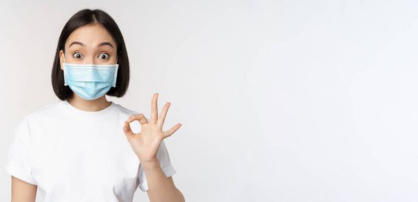 Covid-19, healthcare and medical concept. Impressed asian woman in medical mask, looking amazed and showing ok, okay sign, standing over white background - Zdjęcie, obraz
