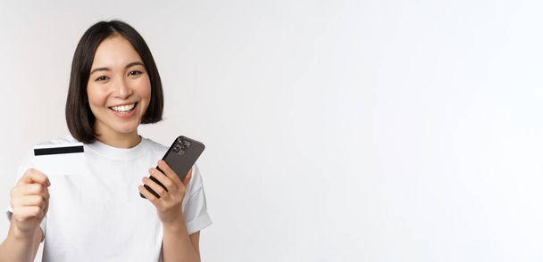 Усміхнена азіатська дівчина використовує мобільний телефон, показує кредитну картку, концепцію безконтактних платежів, онлайн-покупки, стоїть на білому тлі
 - Фото, зображення