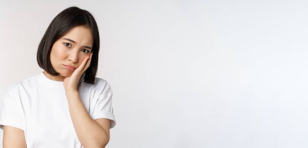 Sad asian girl looking upset and lonely, sulking and frowning, standing against white background in casual tshirt - Photo, Image