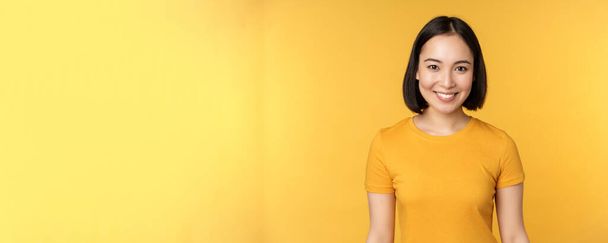 Portré fiatal modern ázsiai nő, mosolygós boldog fehér fogak, magabiztos a kamera, visel alkalmi póló, áll a sárga háttér felett - Fotó, kép