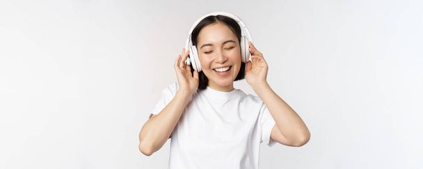 Moderna chica asiática bailando, escuchando música con auriculares, sonriendo feliz, de pie en camiseta sobre fondo blanco - Foto, imagen