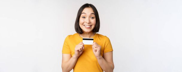 Portrét krásné korejské dívky držící kreditní kartu, doporučujeme bankovní služby, stojící ve žlutém tričku přes bílé pozadí - Fotografie, Obrázek