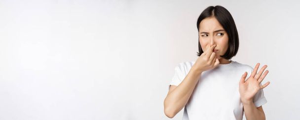 Asian girl looks disgusted, rejecting product with bad smell, shut nose from aversion and cringe, standing against white background - Фото, изображение