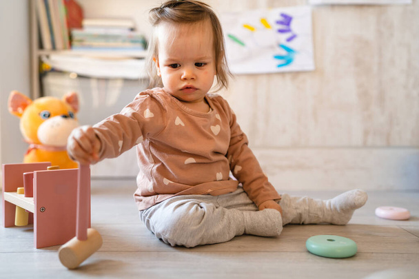 One baby small caucasian infant girl playing on the floor at home copy space holding wooden toys in day front view - Photo, Image