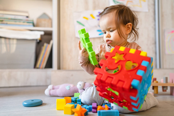 家のコピースペースで床の上で遊んでいる1人の小さな白人の幼児の女の子昼間のフロントビューでプラスチック製のおもちゃを保持 - 写真・画像