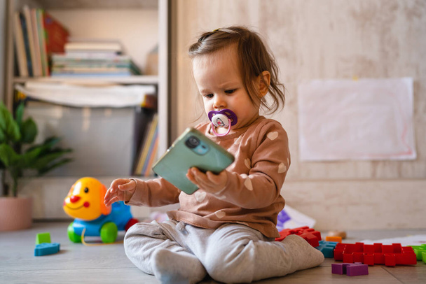 One baby small caucasian infant girl playing on the floor at home copy space holding mobile phone smartphone using to call or watch video in day front view - Photo, Image