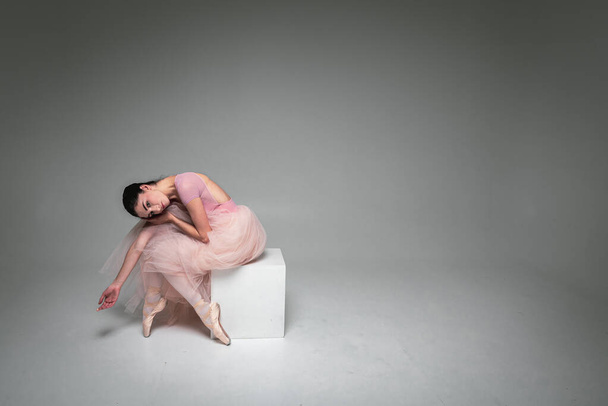 молодая красивая, хрупкая, красивая балерина сидит уставшая, измученная в длинном бледно-розовом платье с тюлем на однородном фоне, движениями рук, сдержанным тоном. Балет, танец, танцор. - Фото, изображение
