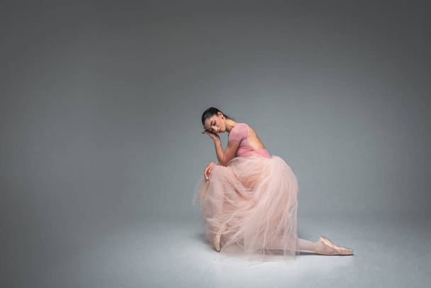 молодая красивая, хрупкая, красивая балерина сидит уставшая, измученная в длинном бледно-розовом платье с тюлем на однородном фоне, движениями рук, сдержанным тоном. Балет, танец, танцор. - Фото, изображение