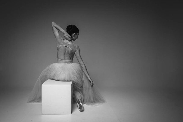 czarno-białe zdjęcie, młoda ładna, krucha, piękna balerina siedząca na białym kwadracie, odwrócone podniesione ręce w długiej blado różowej sukience z tiulu na jednolitym tle, powściągliwy ton. Balet - Zdjęcie, obraz