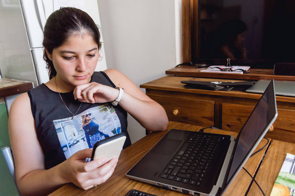 Bělošská mladá žena ve své domácí kanceláři kontroluje vzkazy po telefonu, zatímco pracuje na počítači. sezení u dřevěného stolu - Fotografie, Obrázek