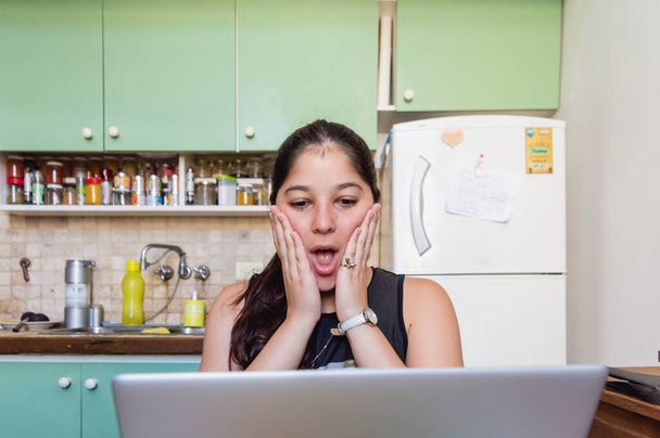 mladá běloška venezuelka doma dívá na písemku na počítači překvapená, že tomu nemůže uvěřit, sedí v obývacím pokoji domu a kontroluje e-maily od fakulty - Fotografie, Obrázek