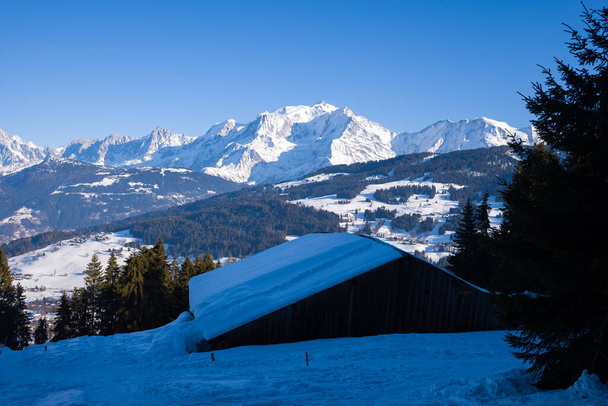 Αυτή η φωτογραφία τοπίου τραβήχτηκε στην Ευρώπη, στη Γαλλία, στο Rhone Alpes, στη Σαβοΐα, στις Άλπεις, το χειμώνα. Βλέπουμε ένα ξύλινο σαλέ απέναντι από το βουνό Mont Blanc, κάτω από τον ήλιο. - Φωτογραφία, εικόνα