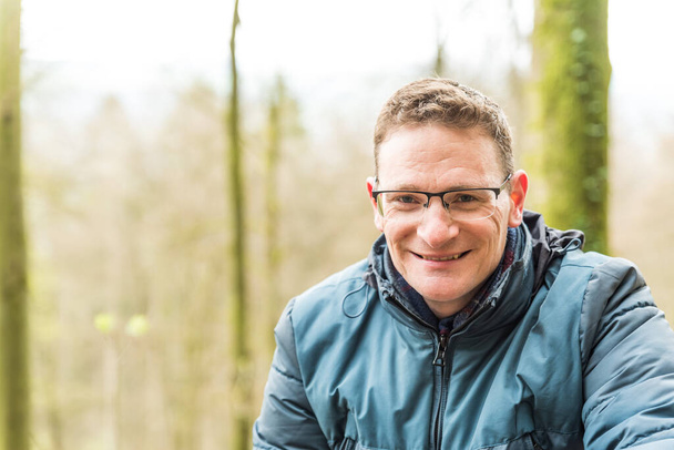 Kordel, Rhénanie-Palatinat - Allemagne - 04 13 2019 - Gros plan portrait d'un homme blanc souriant de quarante ans avec des lunettes et une veste bleue mi-saison avec un fond bokeh vert - Photo, image