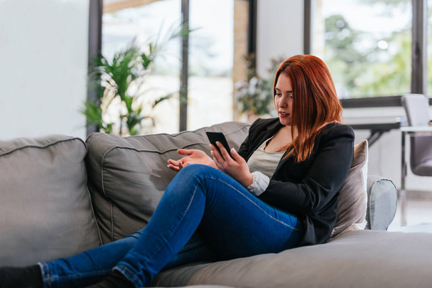 Young woman talking on mobile phone on speakerphone while resting on sofa - Photo, image