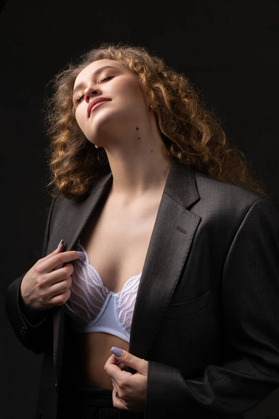 Portrait of a young red-haired curly sexy girl in a jacket with long hair in the studio. Dramatic photo in dark colors. - Fotó, kép