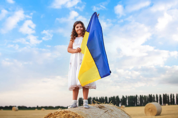少女は地面の上にウクライナ国旗を掲揚した。子供は青い空に対してウクライナの旗を保持しています。ロシアの日に勝利. - 写真・画像