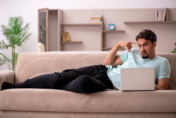 Молодой человек сидит на диване с компьютером дома - Фото, изображение