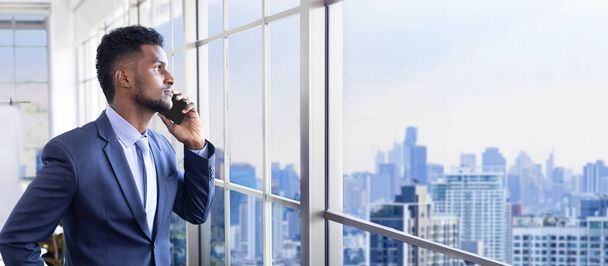 Afrikai-amerikai üzletember hivatalos öltönyben mobiltelefonnal, miközben kinézett az ablakon, hogy a városkép felhőkarcoló kilátás látás, álom és sikeres üzleti és befektetési tervezés - Fotó, kép