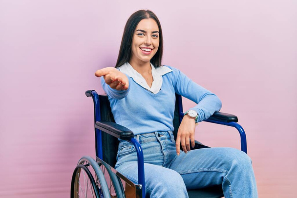 Hermosa mujer con ojos azules sentada en silla de ruedas sonriente alegre ofreciendo la palma de la mano dando asistencia y aceptación.  - Foto, imagen